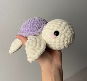 Small crochet amigurumi turtle made with purple and white plush velvet yarn, featuring safety eyes and pink embroidered cheeks.