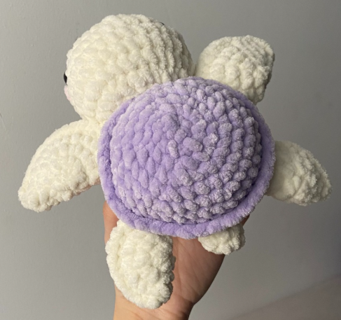 Small crochet amigurumi turtle made with purple and white plush velvet yarn, featuring safety eyes and pink embroidered cheeks.