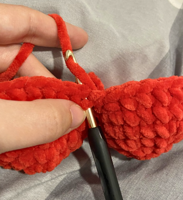 Hands crocheting a red amigurumi heart project with a 4.5 mm crochet hook, working on the upper lobes and joining stitches.