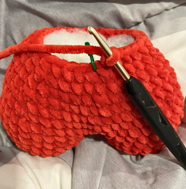 Hands crocheting a red amigurumi heart project with a 4.5 mm crochet hook, working on the upper lobes and joining stitches.