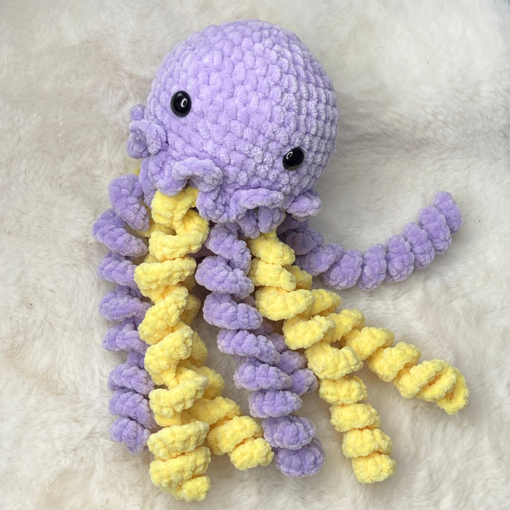 Handmade no-sew crochet jellyfish plushie in purple and yellow yarn with safety eyes and textured curly tentacles, sitting on a neutral background.