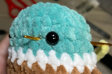Embroidering yellow yarn beak detail on green crochet duck head using a tapestry needle between safety eyes.
