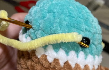 Embroidering yellow yarn beak detail on green crochet duck head using a tapestry needle between safety eyes.