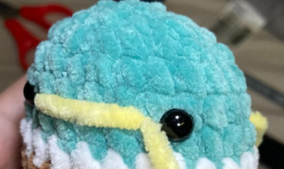 Embroidering yellow yarn beak detail on green crochet duck head using a tapestry needle between safety eyes.