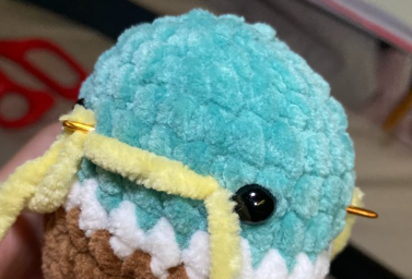 Embroidering yellow yarn beak detail on green crochet duck head using a tapestry needle between safety eyes.